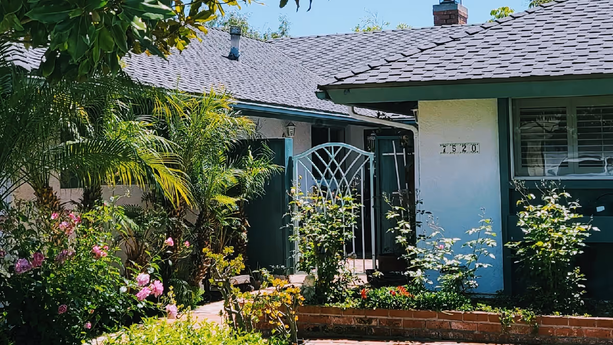 Front exterior view of a single-story residential building with a gray shingled roof, white walls, and green trim. The entrance features a decorative metal gate surrounded by lush greenery, including palm plants and flowering bushes with pink blooms. The house number 7520 is visible on the wall near a window with closed blinds.