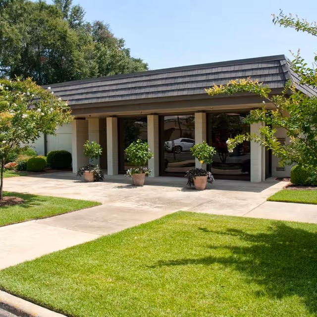 Exterior view of a single-story building with a dark shingled roof and large glass windows. There are four potted plants placed in front of the building, surrounded by well-maintained green grass and small trees on either side. The sky is clear and the scene is bright and sunny.