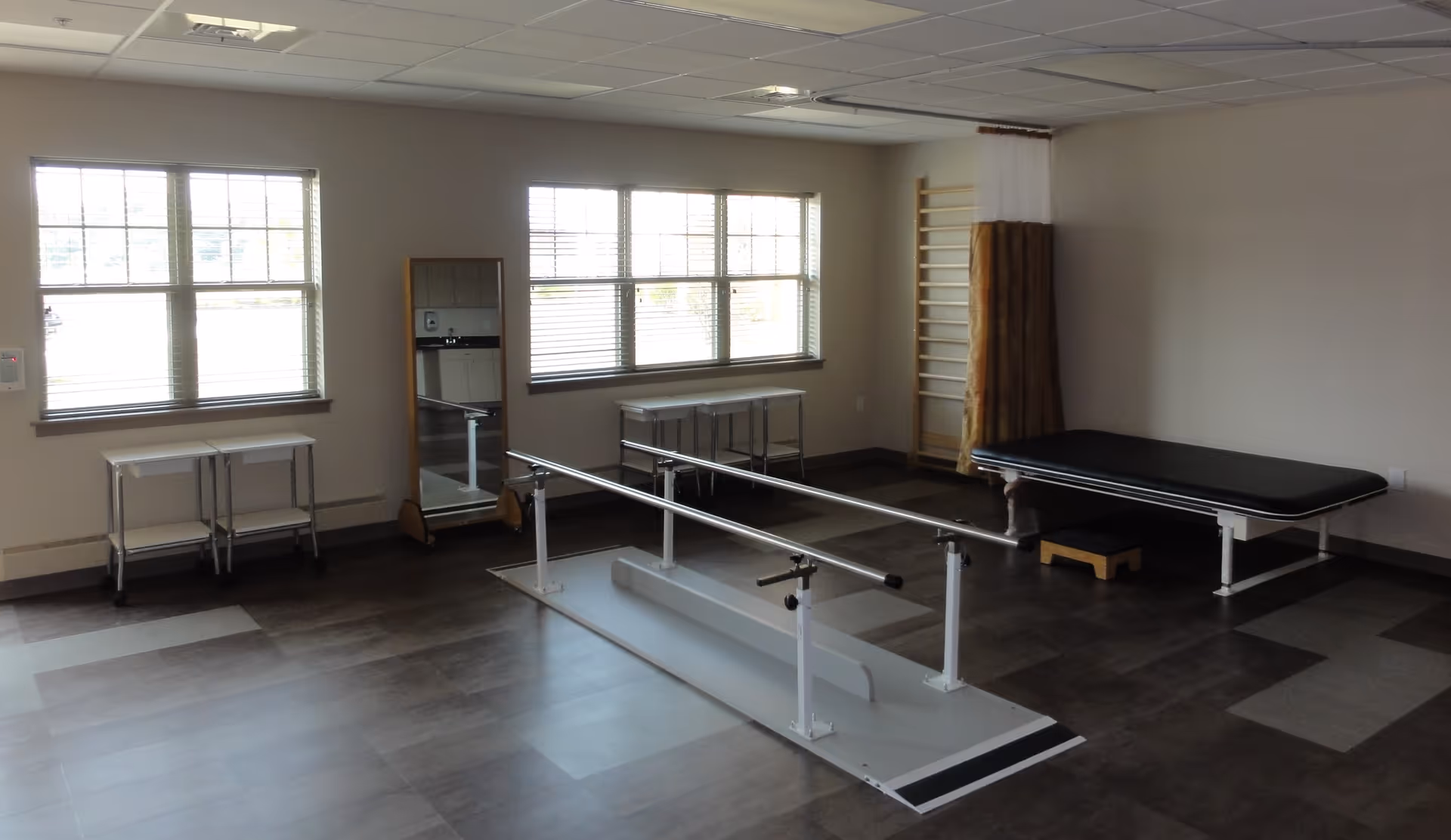 Empty physical therapy room with parallel walking bars, an exam table, and windows.