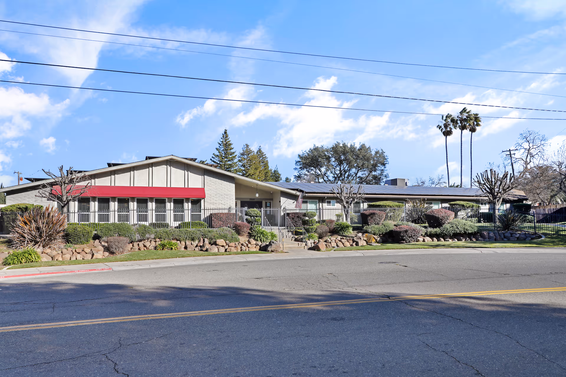Single-story assisted living building with a red awning, landscaped shrubs, and a fenced front yard facing a street.