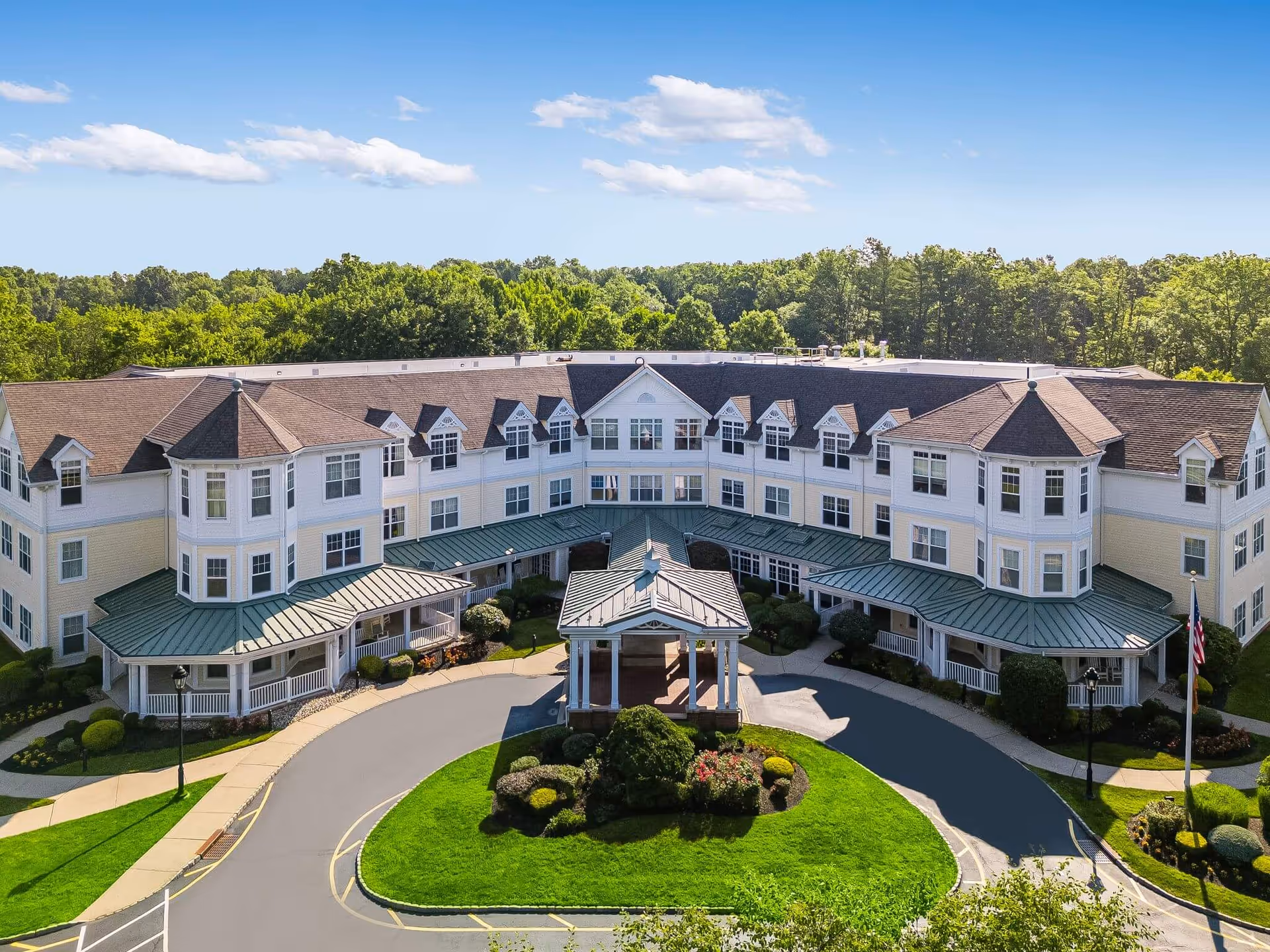 A large, three-story senior living facility building with white and light yellow exterior walls, green metal awnings, and multiple windows. The building is U-shaped with a covered entrance in the center, surrounded by well-maintained landscaping and a circular driveway. Trees and a clear blue sky are visible in the background.