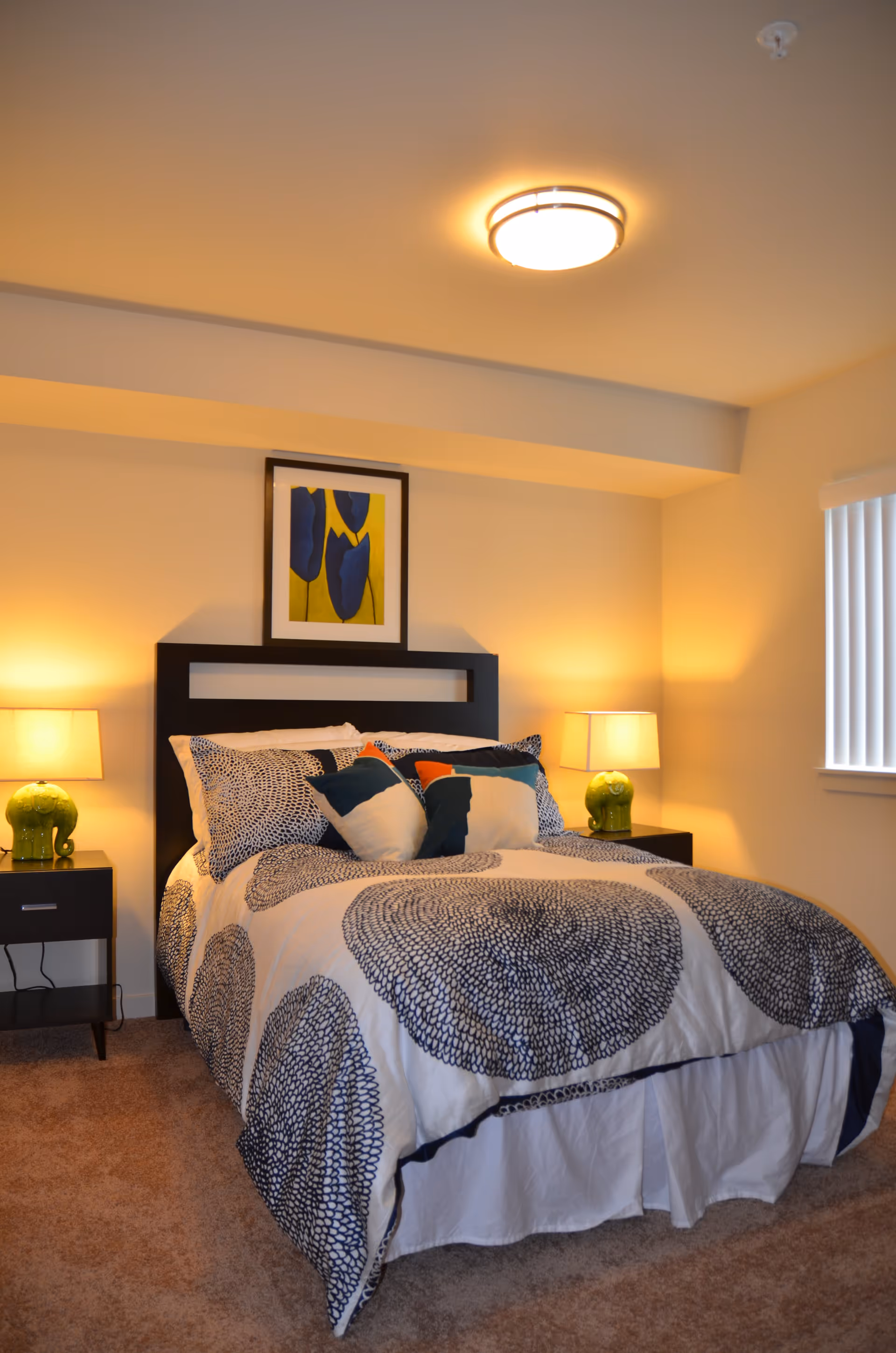 A cozy bedroom with a bed featuring a white and navy blue patterned comforter and multiple pillows. There are two black nightstands on either side of the bed, each with a green elephant-shaped lamp. A framed artwork with blue and yellow colors hangs above the bed. The room has beige carpet and walls, a ceiling light fixture, and a window with vertical blinds.
