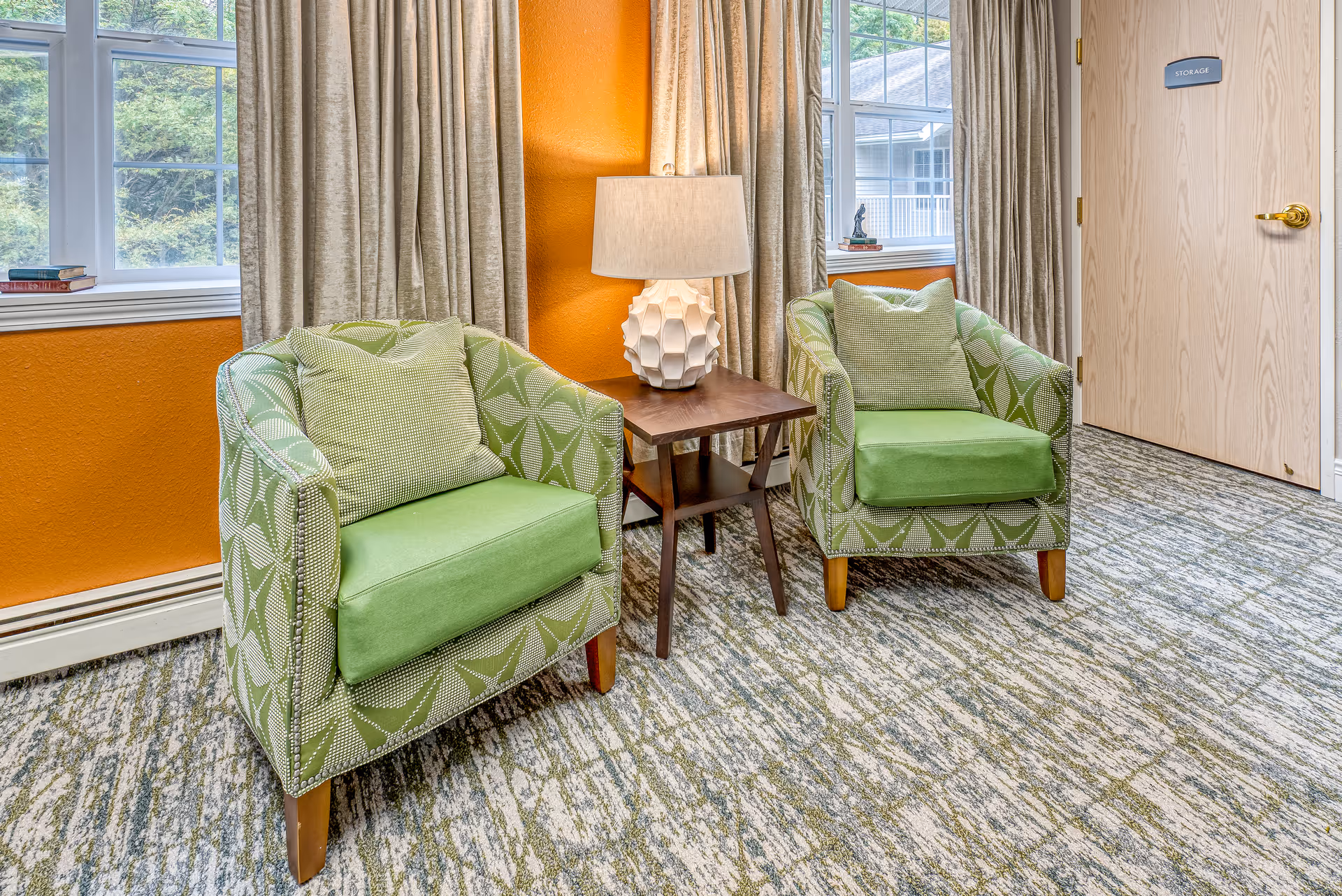 A cozy seating area with two green patterned armchairs, each with a green cushion, positioned on a patterned carpet. Between the chairs is a wooden side table with a white textured lamp. The background features orange walls, large windows with beige curtains, and a wooden door labeled 'STORAGE'.