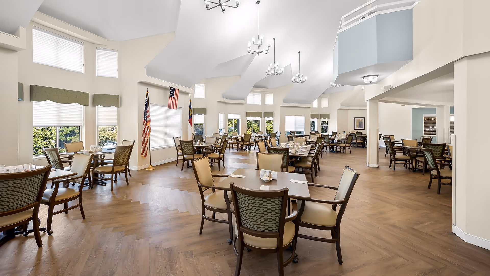 A spacious dining room with multiple tables and chairs arranged neatly. The room has large windows with white blinds and green valances, allowing natural light to brighten the space. There are two flags displayed near the windows, and modern chandeliers hang from the high ceiling. The floor is wooden with a herringbone pattern, and the overall atmosphere is clean and inviting.