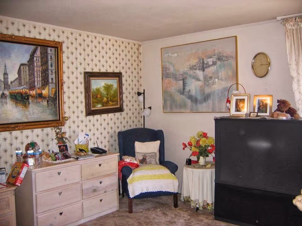 A cozy retirement living room with patterned wallpaper, framed paintings, a blue armchair with a knit throw, a dresser, a round side table with flowers, and a large black television.