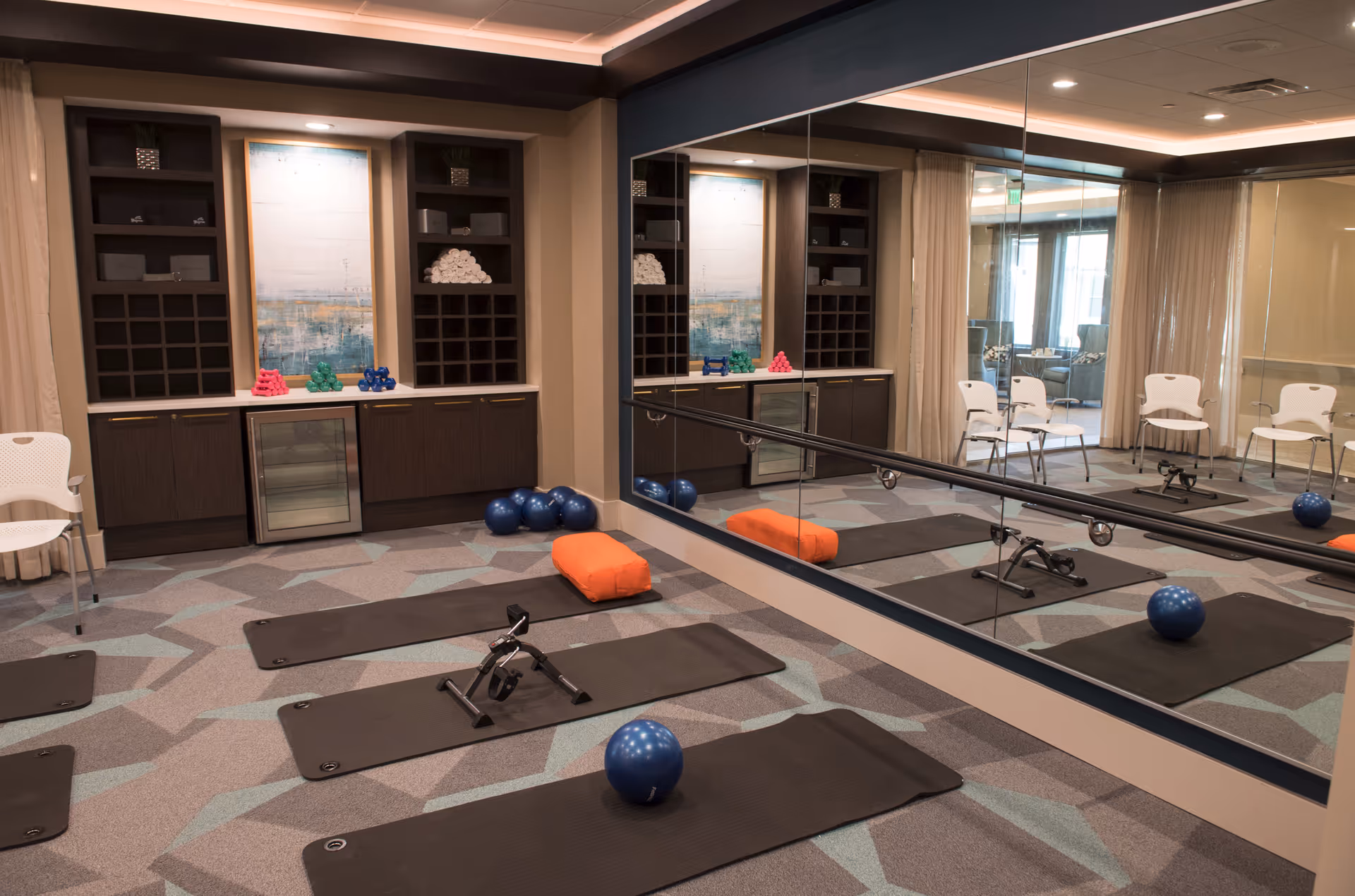 Exercise room with yoga mats, small exercise bikes, blue exercise balls, and colorful dumbbells. The room has a large mirror on one wall, a cabinet with rolled towels, and a modern abstract painting. There are also white chairs along the wall and a patterned carpet.