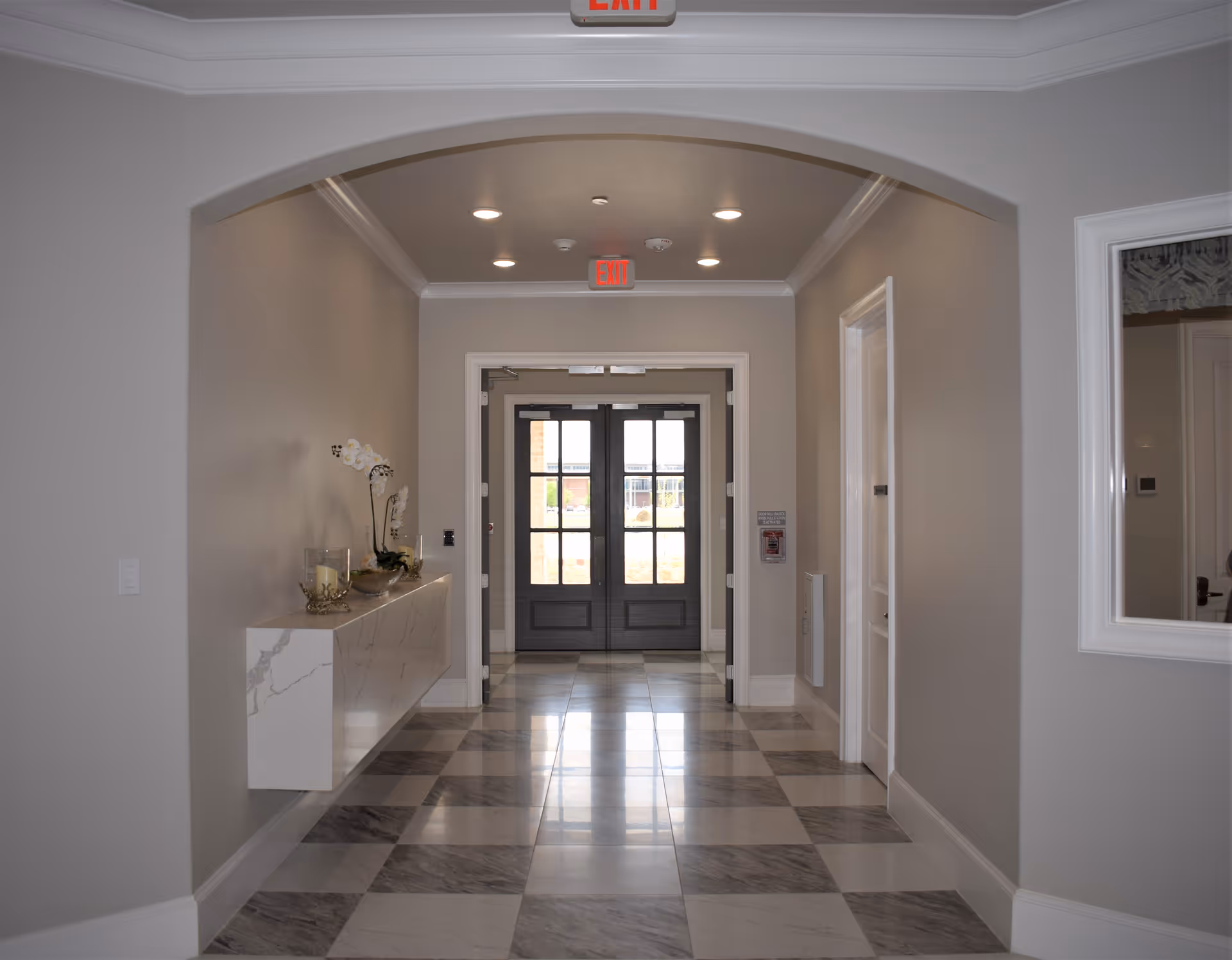 A clean, well-lit hallway with a checkered marble floor leading to double glass doors. The hallway has light gray walls, a white ceiling with recessed lighting, and an archway. A floating marble shelf on the left holds decorative items including candles and flowers. An exit sign is visible above the doors.