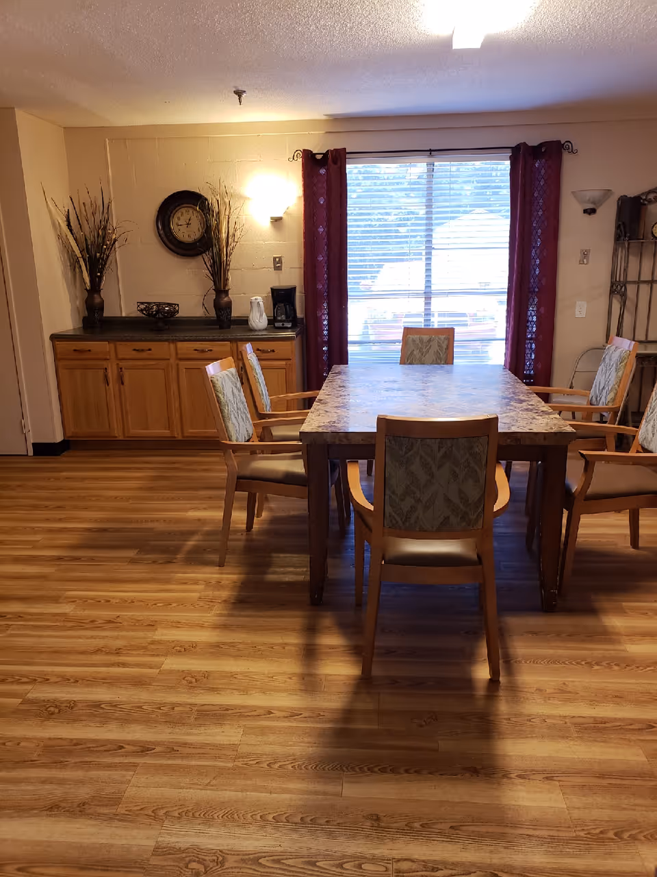 A dining room with a rectangular table and chairs, wooden cabinets against the wall, and a window with red curtains.