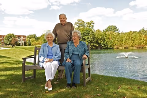 Two elderly women sitting on a wooden bench and an elderly man standing behind them, all smiling, in a grassy outdoor area near a pond with a fountain. Trees and a building are visible in the background under a partly cloudy sky.