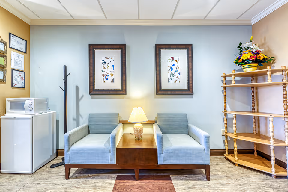 A small seating area with two light blue armchairs separated by a wooden side table with a lamp. Behind the chairs are two framed floral artworks on a light blue wall. To the left, there is a white mini fridge with a microwave on top and a black coat rack. On the right, there is a wooden shelving unit with a colorful flower arrangement on the top shelf. The floor has a wood-like finish.