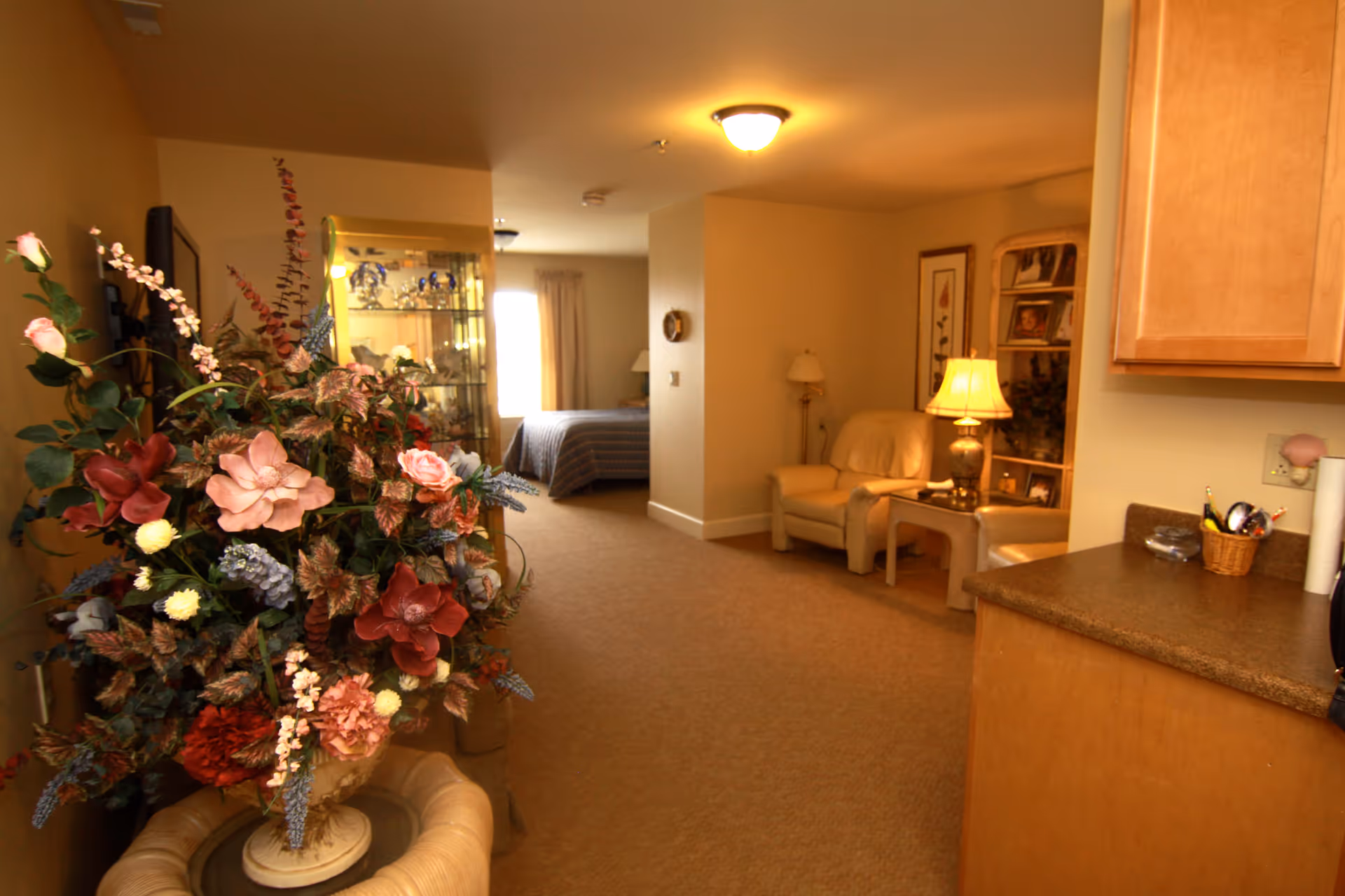 Open interior of a senior living suite with a large floral arrangement in the foreground, a seating area and lamp to the right, a kitchenette counter at the right edge, and a bed visible in the background.