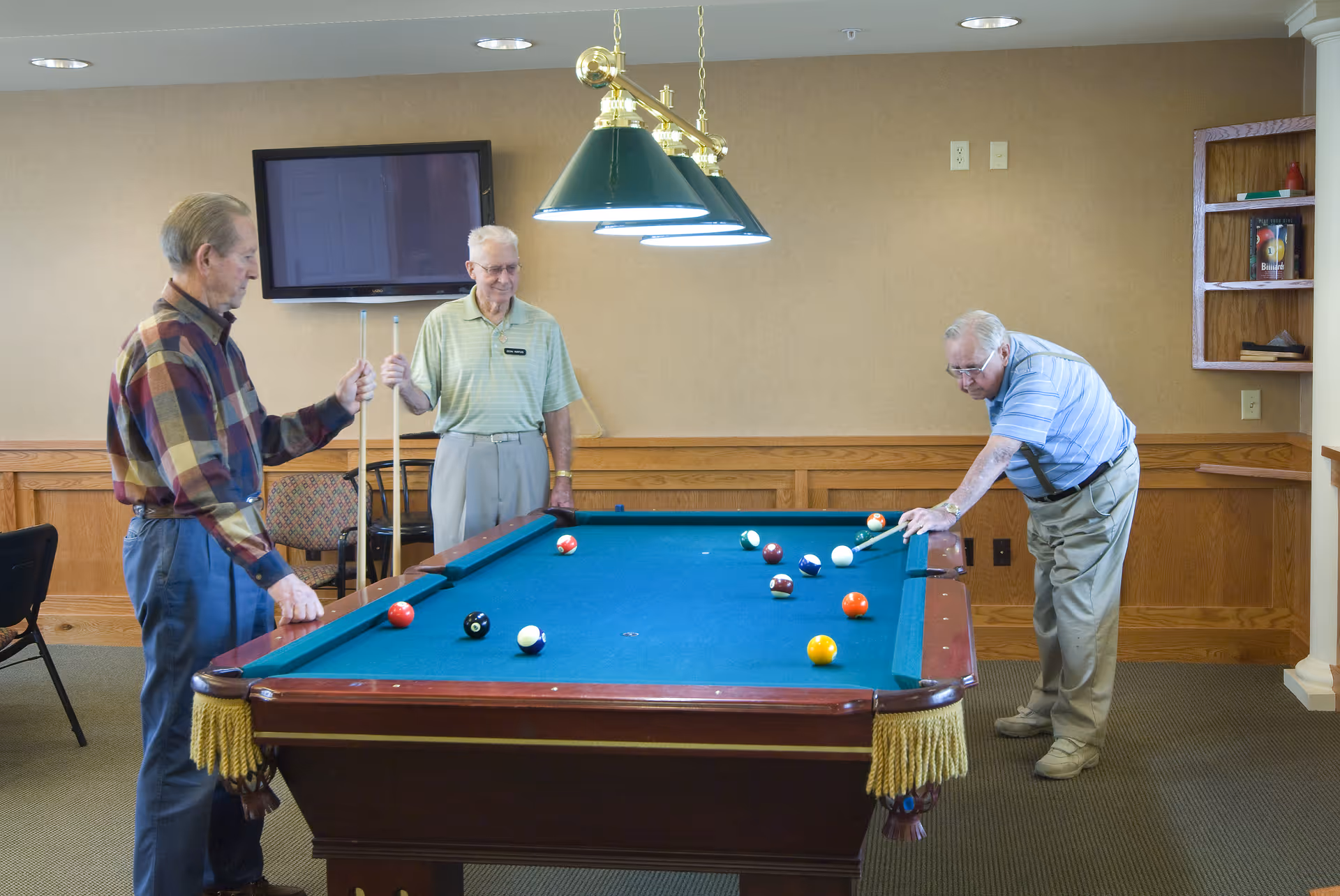 Three elderly men playing pool in a well-lit room with beige walls and wood paneling. One man is taking a shot while the other two watch, each holding a pool cue. A flat-screen TV is mounted on the wall behind them, and a green hanging light fixture illuminates the pool table.