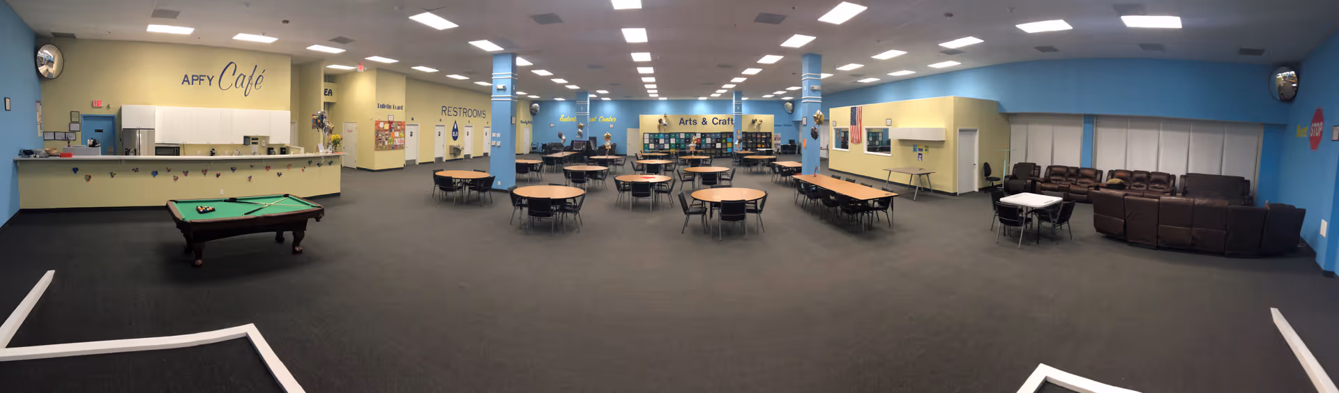 Wide panoramic view of a large indoor common area in a senior living facility. The room features a pool table, multiple round tables with chairs, a café counter labeled 'APFY Café', an arts and crafts section with shelves and supplies, a seating area with brown leather couches, and signage indicating restrooms and other facility areas. The walls are painted light blue and yellow, and the space is well-lit with ceiling lights.