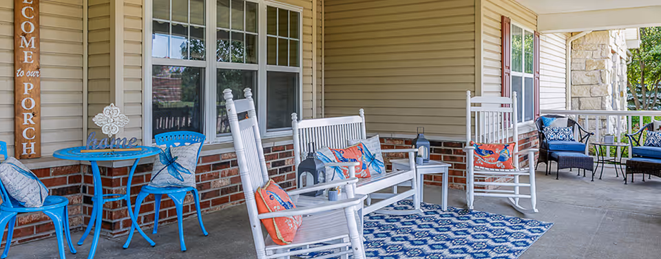 A covered porch area with white rocking chairs and a bench adorned with colorful pillows, a blue patterned rug, a small blue table with two blue chairs, and decorative items including a wooden sign that reads 'Welcome to our Porch'. The porch has beige siding, brick accents, and windows, with greenery visible outside.