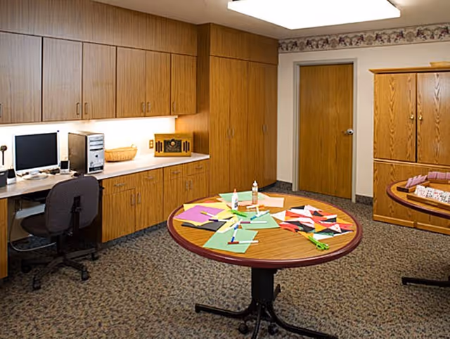 A room with wood-paneled cabinets and a computer workstation on the left side. Two round tables in the center and right side of the room have colorful paper, glue, scissors, and craft supplies spread out on them. The room has carpeted flooring, a closed wooden door, and a wooden cabinet against the far wall.