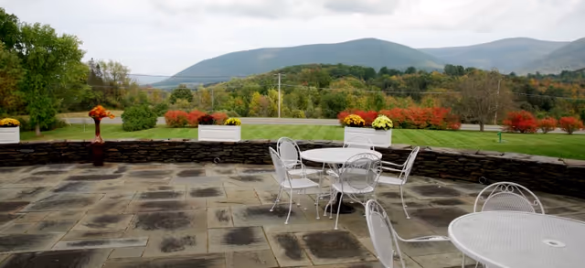 Outdoor patio area with stone flooring and white metal tables and chairs. There are flower planters with yellow and orange flowers placed on a low stone wall. In the background, there is a green lawn, trees with autumn foliage, and rolling hills under a cloudy sky.
