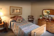 A cozy bedroom in a senior living facility featuring a single bed with a patterned bedspread and multiple pillows. Next to the bed is a wooden nightstand with a lamp and a telephone. On the wall above the bed is a framed artwork. Across from the bed is a wooden dresser with a large mirror, decorative items, and a plant. A wooden chair is placed against the wall near the dresser.