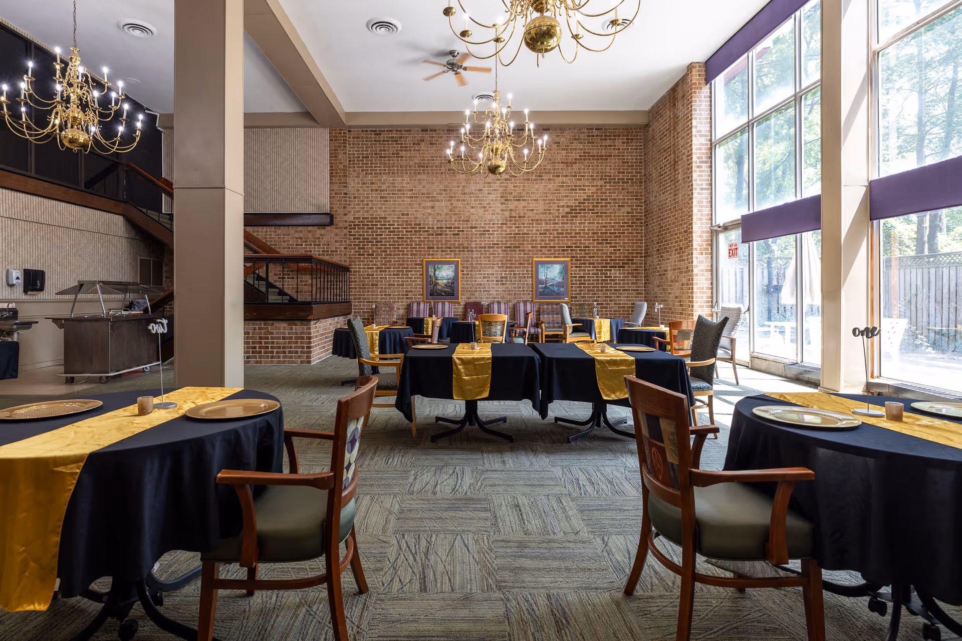 A spacious dining room with large floor-to-ceiling windows on the right side letting in natural light. The room features multiple tables covered with black tablecloths and gold runners, set with plates and napkins. Wooden chairs with cushioned seats surround the tables. The back wall is exposed brick with two framed paintings. Two ornate chandeliers hang from the ceiling, and a staircase is visible on the left side of the room.