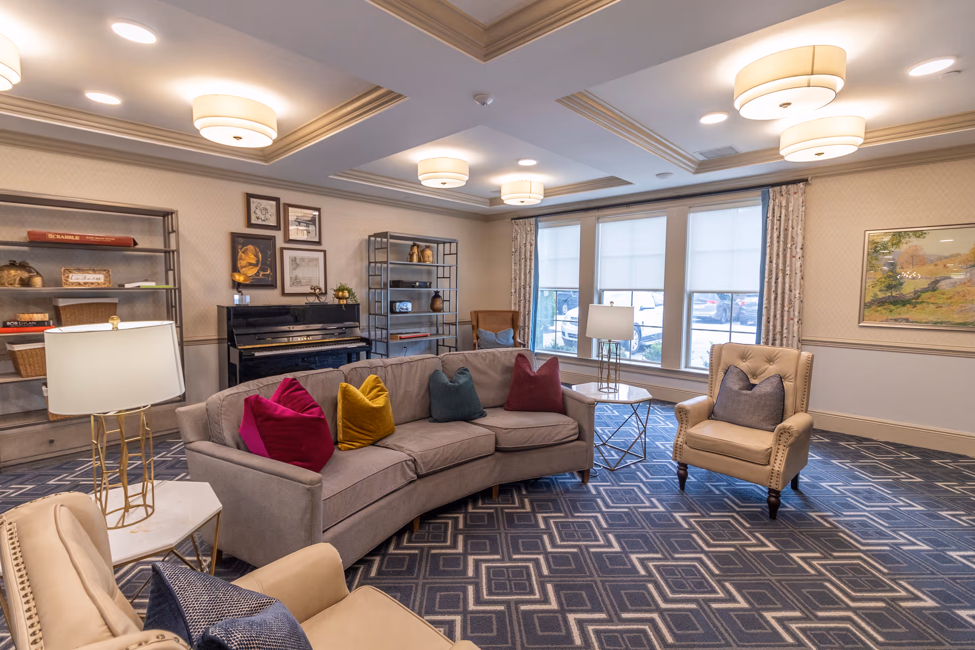 A cozy living room area in a senior living facility featuring a curved beige sofa with colorful throw pillows, two beige armchairs with cushions, a piano against the wall, two metal shelving units with decorative items, large windows with curtains, and modern ceiling lights.