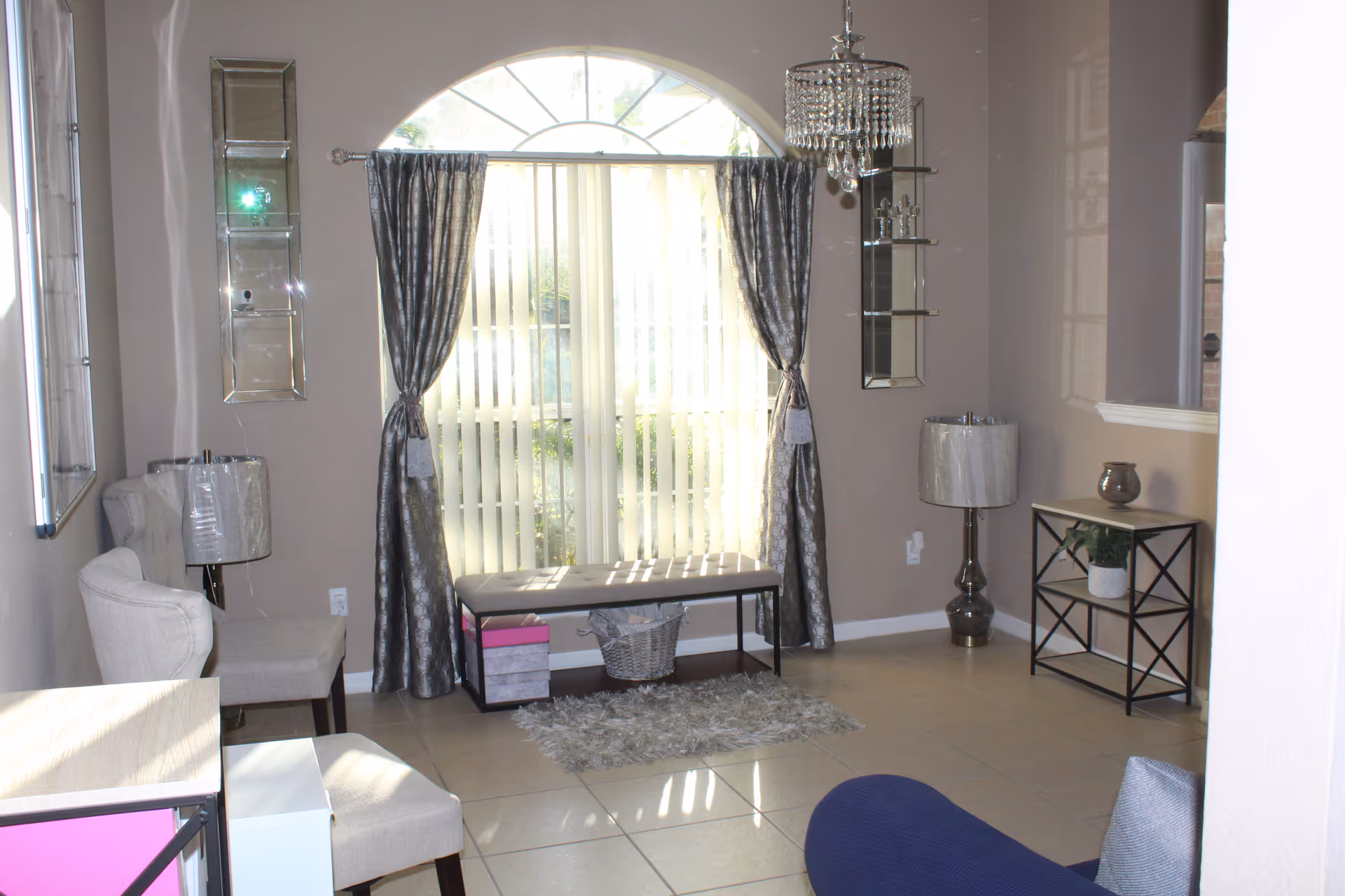 A bright living room with a large arched window covered by vertical blinds and silver curtains. The room features a bench with storage baskets underneath, two beige upholstered chairs, a small rug, two table lamps with silver bases and shades, a small black metal and wood shelving unit with a plant and decorative item, and a crystal chandelier hanging from the ceiling.