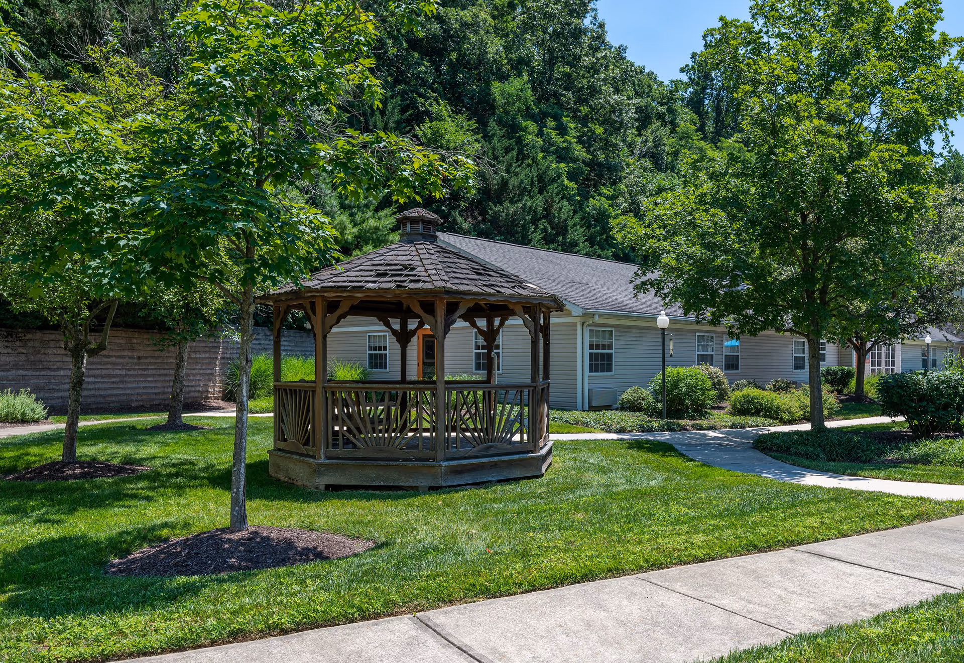 A wooden gazebo with a shingled roof sits on a well-maintained grassy area surrounded by trees and shrubs. Behind the gazebo, there is a single-story building with beige siding and white-framed windows. A concrete walkway curves around the gazebo and leads to the building. The scene is bright and sunny with a clear blue sky.