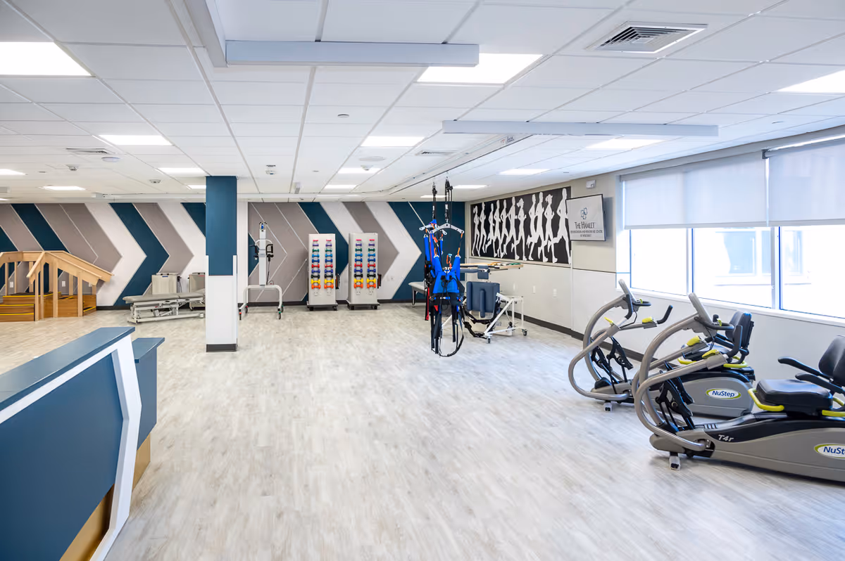 A spacious rehabilitation and exercise room with light wood flooring and a white ceiling. The room features exercise equipment including NuStep machines on the right, colorful dumbbells on racks in the back, parallel bars, and a blue harness system hanging from the ceiling. The walls have a geometric pattern in blue, gray, and white, and a mural of walking silhouettes. Large windows on the right side allow natural light to enter. A sign on the wall reads 'The Hamlet Rehabilitation and Healthcare Center at Nesconset.'