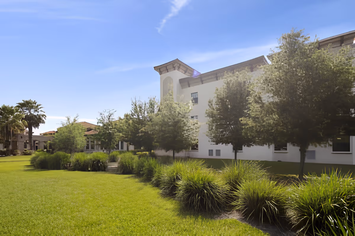 Green lawn and landscaped shrubs and trees in front of a white multi-story senior living building under a clear blue sky.