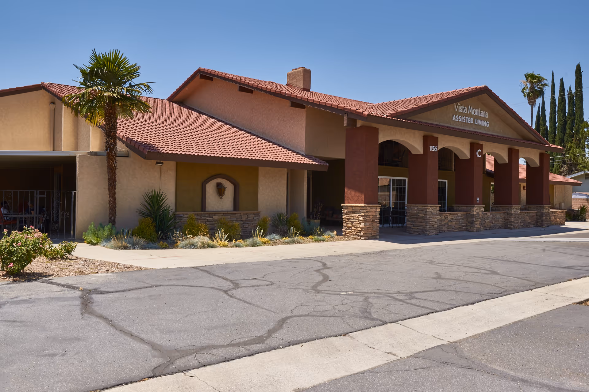 Exterior view of Vista Montana Assisted Living facility with a beige and brown building featuring a red tiled roof, a covered entrance supported by stone pillars, and desert landscaping including palm trees and shrubs under a clear blue sky.