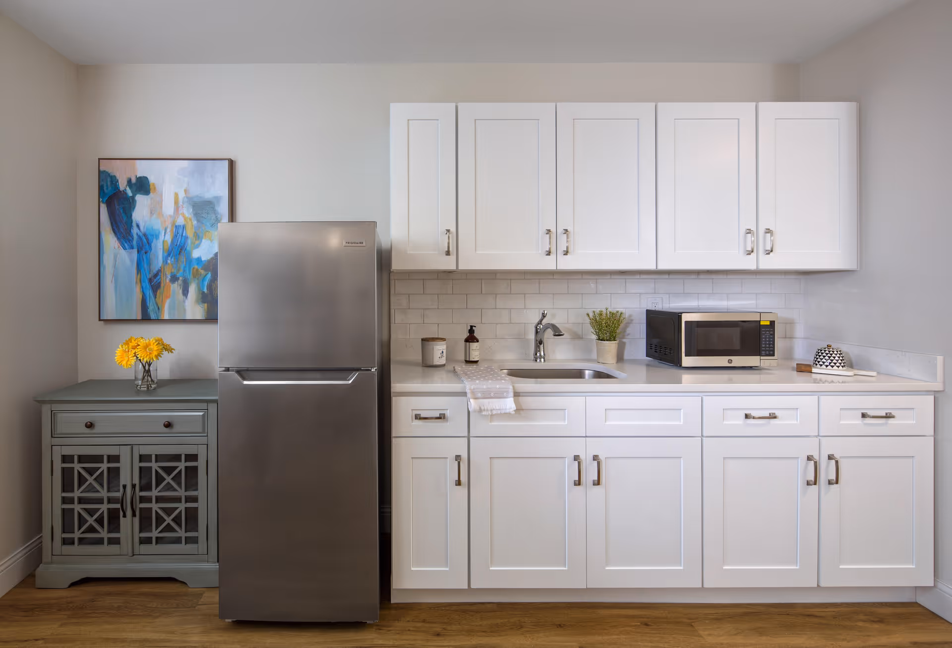 A modern kitchen area with white cabinets and drawers, a stainless steel refrigerator, a sink with a faucet, a microwave, and a small potted plant on the countertop. To the left of the refrigerator is a small green cabinet with a vase of yellow flowers on top and an abstract painting hanging on the wall above it. The floor is wooden.