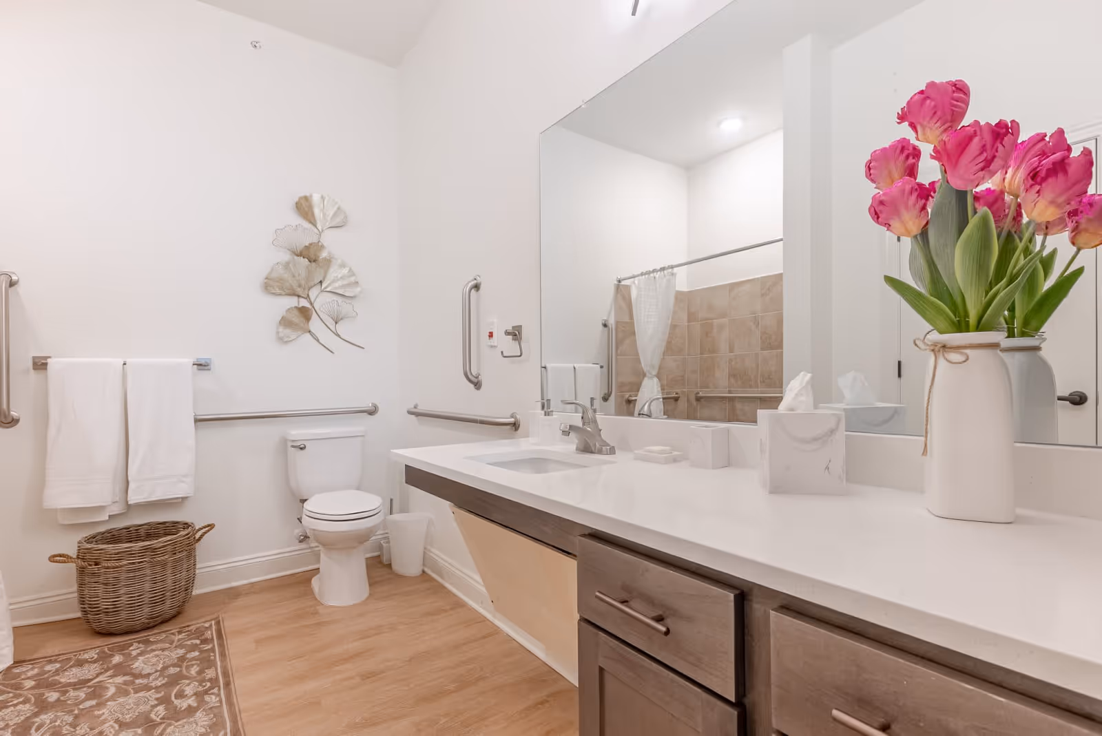 A clean and well-lit bathroom with a white toilet, a large mirror above a countertop with a sink, a vase with pink flowers, a tissue box, and a soap dish. The bathroom has wooden flooring, a wicker basket, white towels hanging on a rail, and a decorative metal wall art piece. There are grab bars installed near the toilet and in the shower area, which has beige tiles and a white shower curtain.