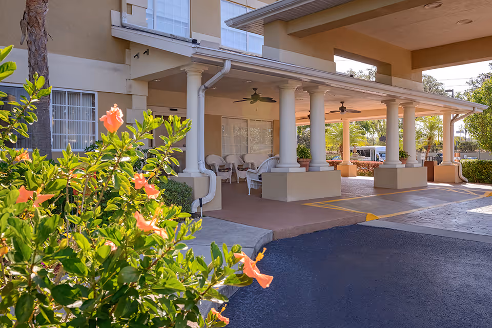 Entrance area of The Bridge at Ocala facility showing a covered driveway with white columns, ceiling fans, and white wicker chairs. There are green plants and orange flowers in the foreground.