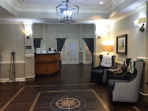 Interior view of a reception area with a wooden front desk, two upholstered armchairs with blankets, a side table with decorative items, wall sconces, framed artwork, and a ceiling light fixture. The floor has a decorative compass design in the center.