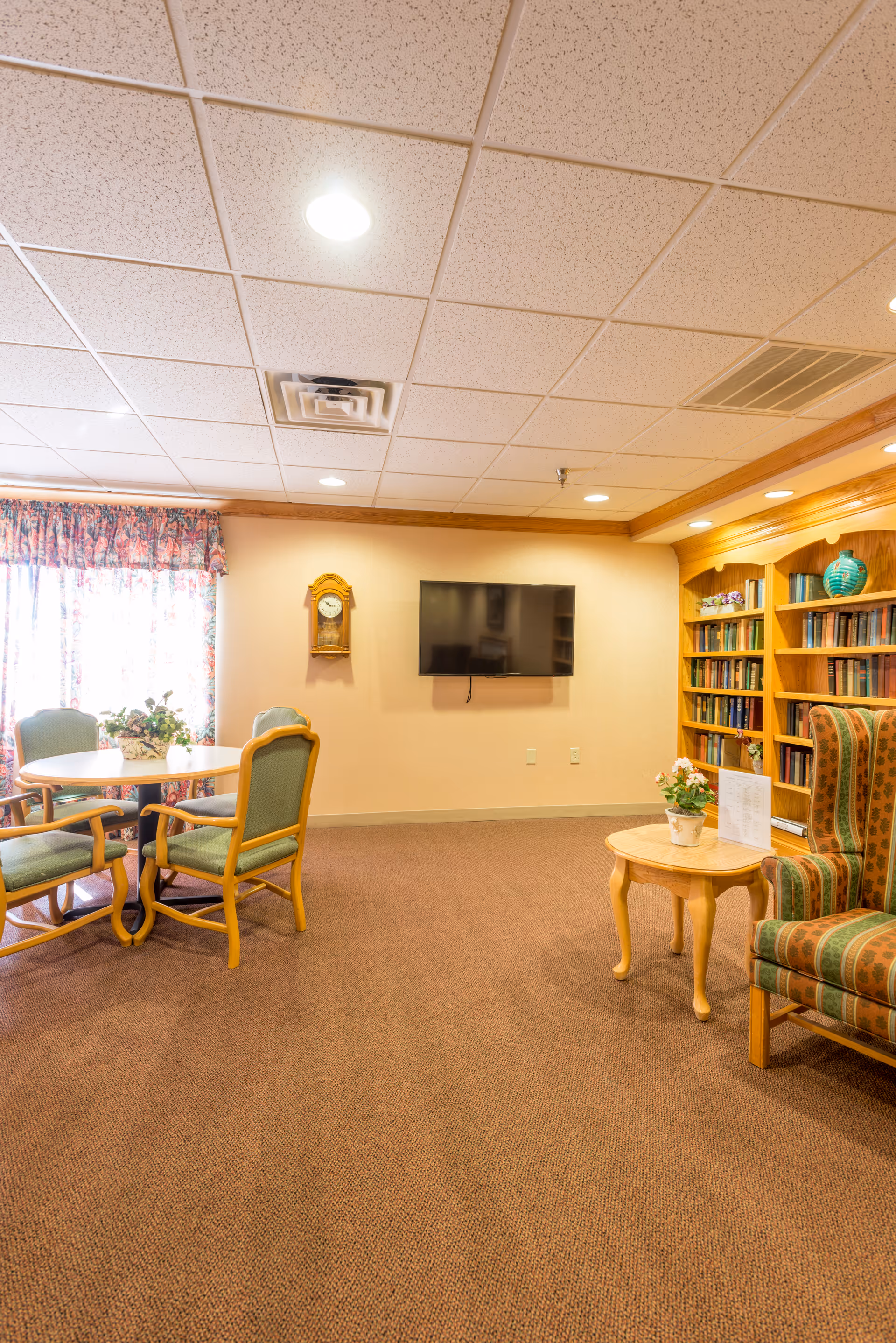 A cozy common area with a round table and four green cushioned chairs near a window with floral curtains. A wall-mounted flat screen TV and a wooden clock are on the beige wall. To the right, there is a wooden bookshelf filled with books and decorative items, and an upholstered armchair with a small wooden side table holding a potted plant and a menu or pamphlet.