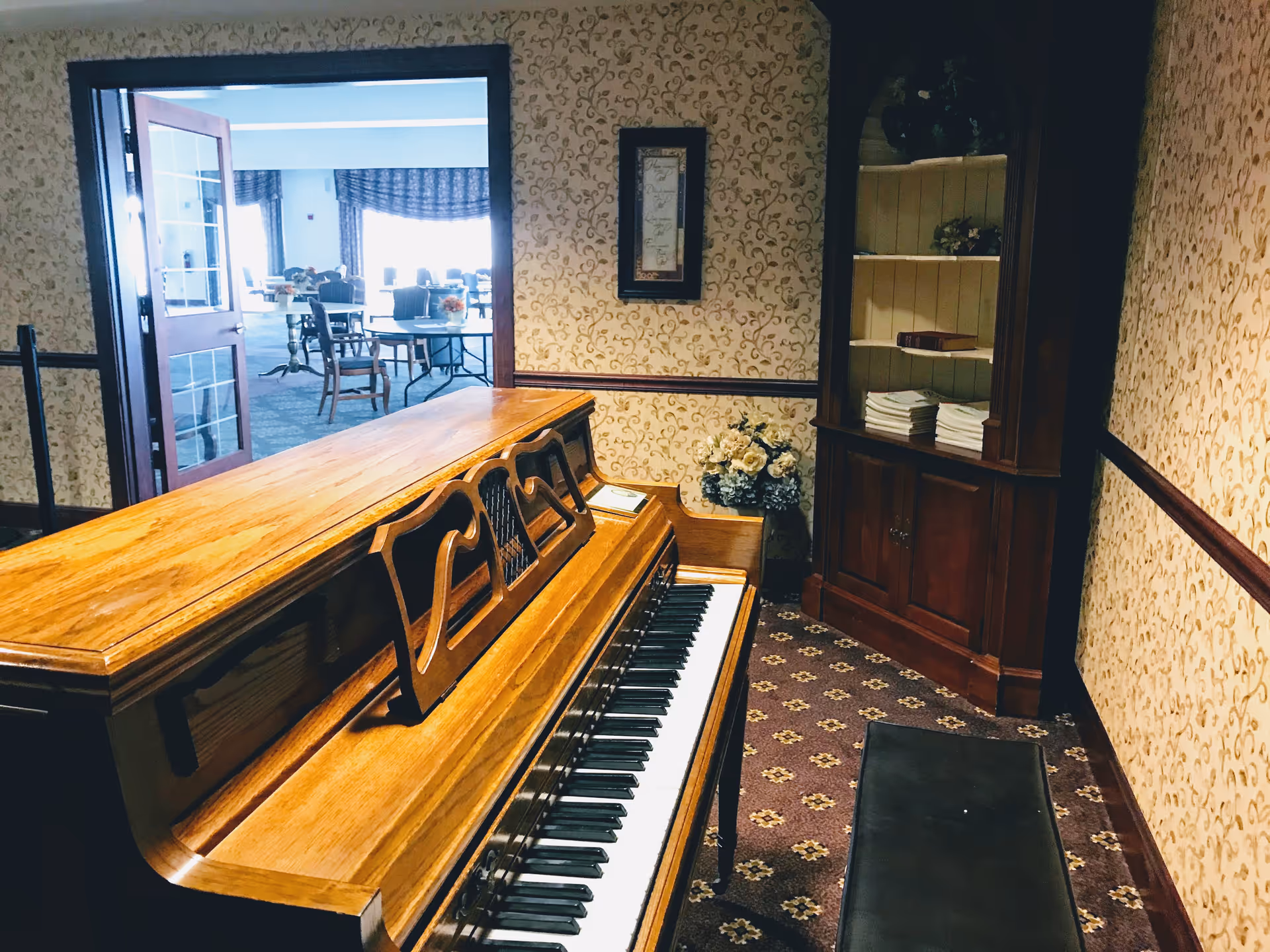 A wooden upright piano with a matching bench in a room with patterned wallpaper and carpet. To the right is a wooden bookshelf with books and decorative items. An open door leads to a bright room with tables and chairs, possibly a dining or common area.
