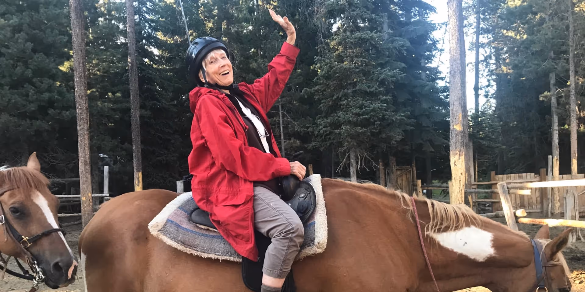 An elderly woman wearing a red jacket and a black helmet is sitting on a brown horse with a white patch on its face, smiling and waving her hand. Another horse is partially visible on the left side. The background shows a wooded area with tall trees and wooden fencing.