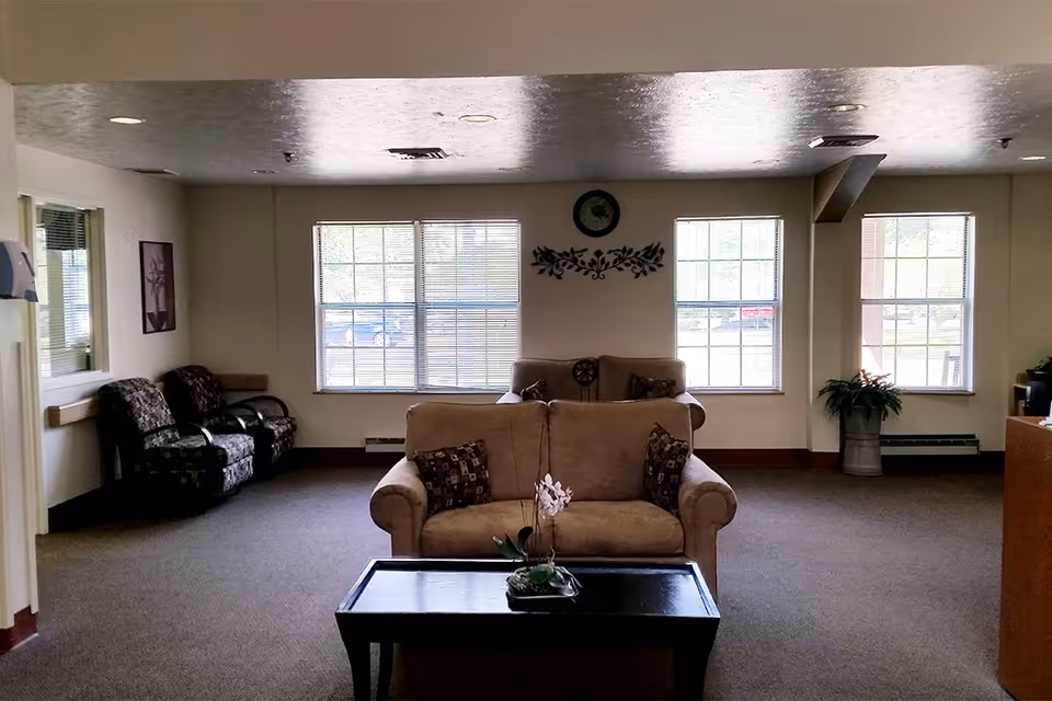 A quiet living room area with a beige loveseat and a dark wooden coffee table with a small plant on it. Behind the loveseat is another beige couch with decorative pillows. The room has three large windows letting in natural light, a wall clock, and a decorative wall piece. To the left, there are two patterned armchairs and a framed picture on the wall. A potted plant is visible near the right window.