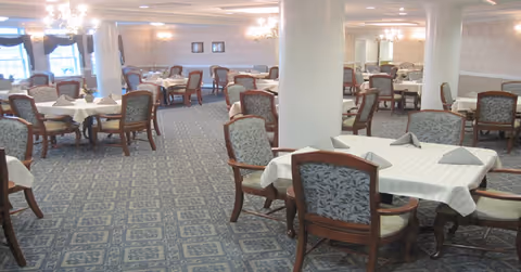 Large carpeted dining room with multiple tables covered in white tablecloths and wooden chairs arranged under chandeliers.