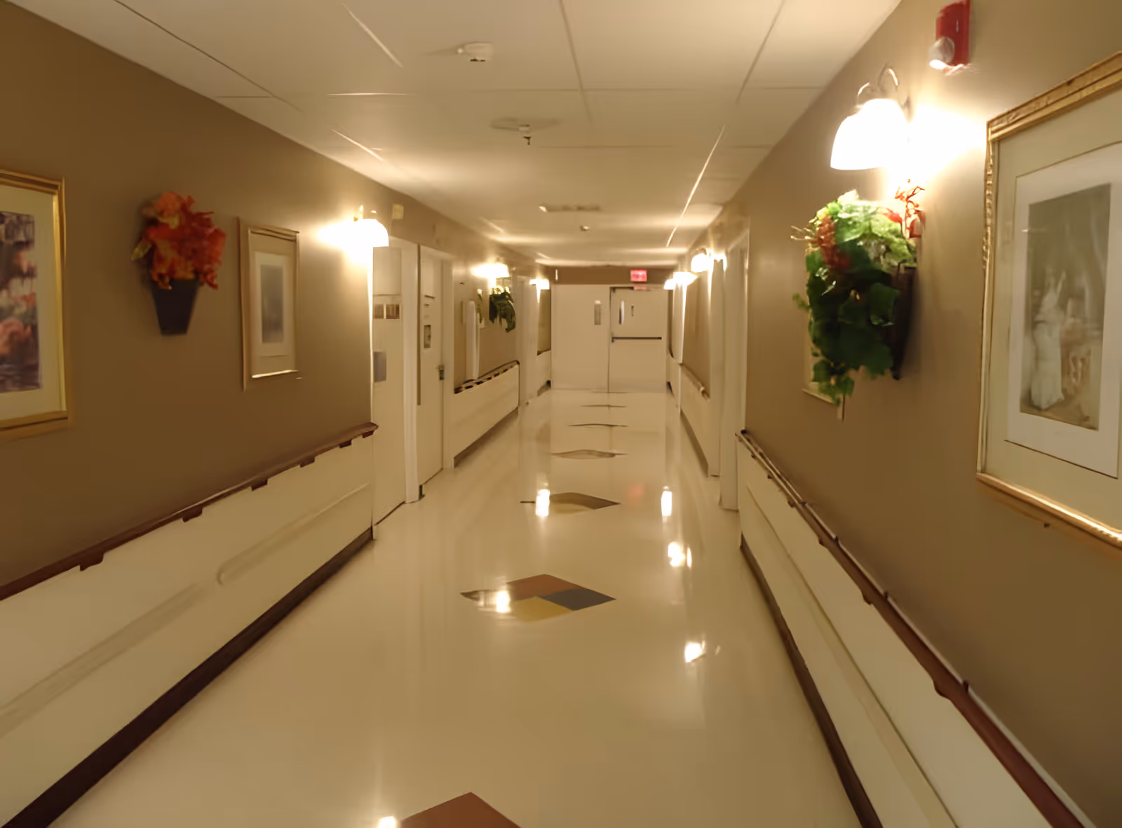A well-lit hallway in a senior living facility with beige walls, handrails on both sides, framed artwork, and decorative wall-mounted flower arrangements. The floor is shiny with diamond-shaped patterns, and double doors are visible at the end of the corridor.