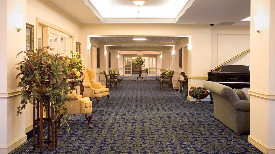 A long, carpeted hallway in a senior living facility with floral patterned blue carpet, beige walls, and soft lighting. The hallway is decorated with potted plants, yellow upholstered chairs, wooden side tables, and framed artwork on the walls. On the right side, there is a black grand piano next to a gray couch and a bronze statue of a child reading a book. At the far end, double glass doors and more floral arrangements are visible.