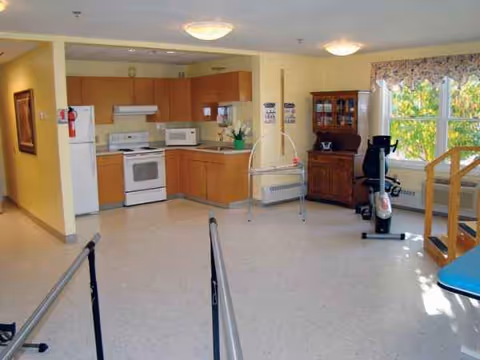 Common living area with kitchenette, exercise equipment, and large windows in a senior residence.