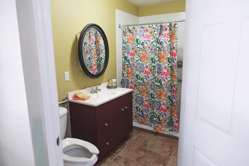 A bathroom with a white toilet, a dark wood vanity with a white sink, a round mirror above the sink, and a shower with a colorful floral shower curtain. The walls are painted light yellow and the floor is tiled.