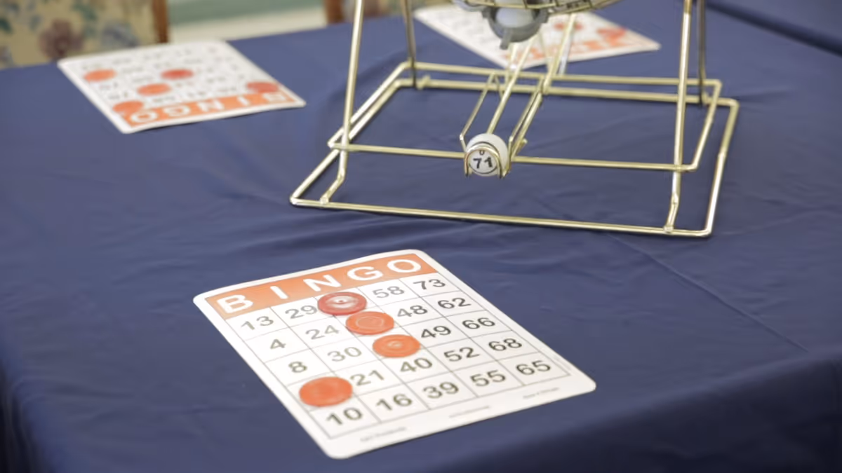 A bingo card on a blue tablecloth with several red markers placed on it, and a metal bingo cage with numbered balls in the background.