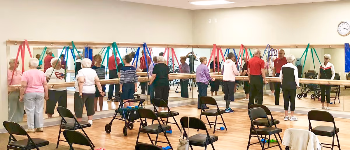 A group of elderly individuals participating in a fitness or physical therapy class in a mirrored exercise room. They are using colorful resistance bands attached to a ballet barre along the wall. Several folding chairs and walkers are placed around the room.