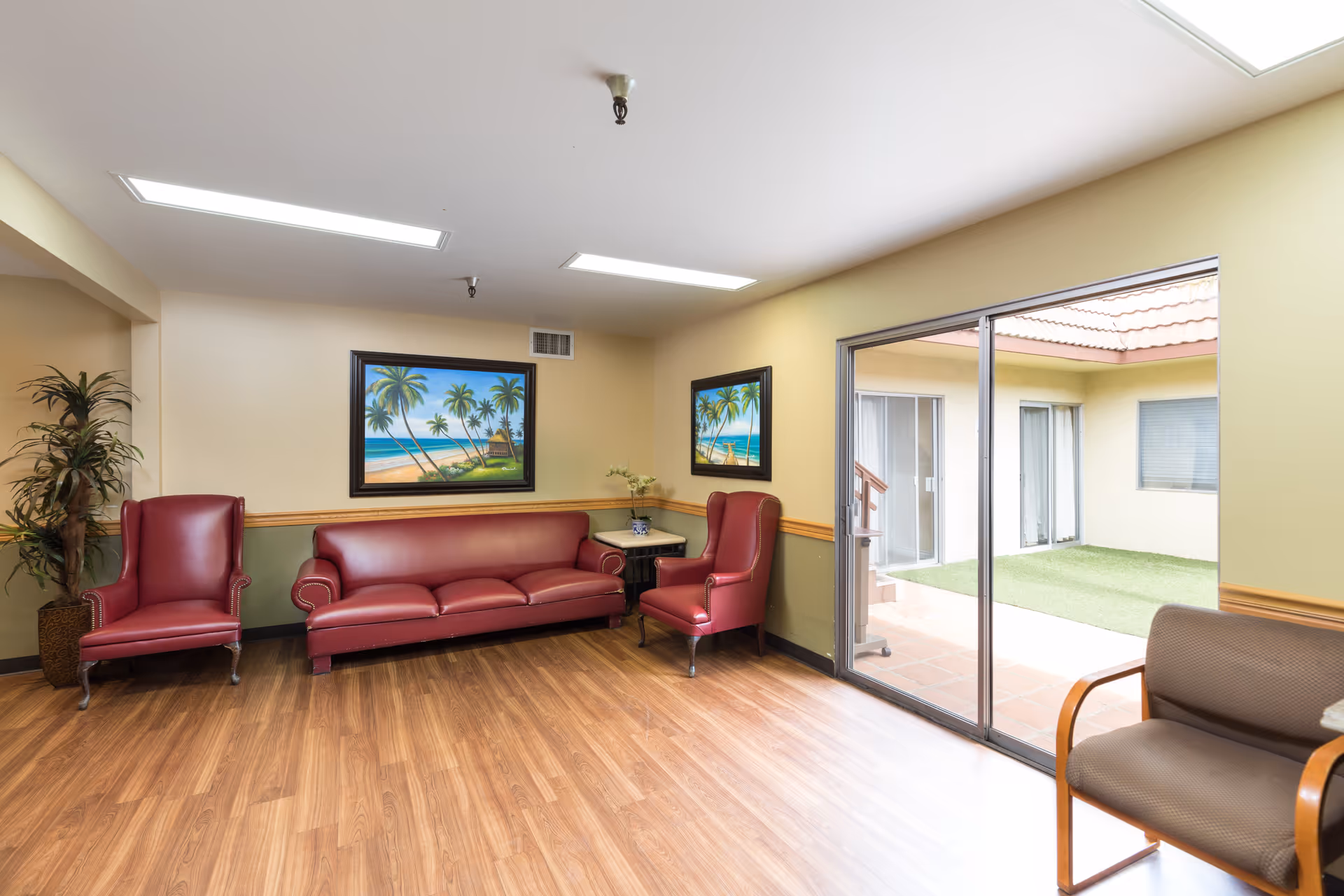 A seating area in a healthcare facility with two red armchairs and a red sofa arranged around a small table. The walls are painted light yellow with a green lower half and decorated with framed paintings of tropical beach scenes. There is a large sliding glass door leading to an outdoor courtyard with artificial grass and a tiled patio. The floor is wood laminate, and there is a potted plant in the corner.