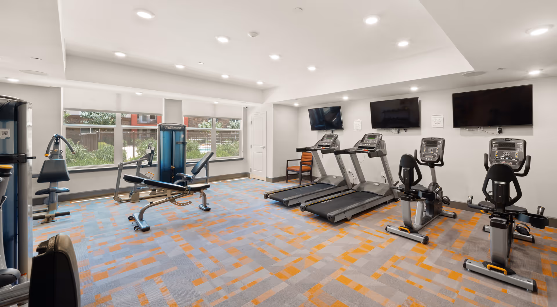 A well-lit fitness room with exercise equipment including two treadmills, two stationary bikes, and a multi-function weight machine. The room has large windows letting in natural light and three wall-mounted flat-screen TVs. The floor is carpeted with a pattern of gray and orange squares.