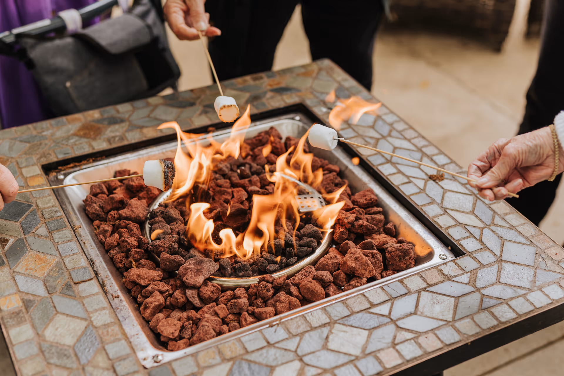 Close-up of three people roasting marshmallows over a square outdoor fire pit with mosaic tile edges and lava rocks surrounding the flames.
