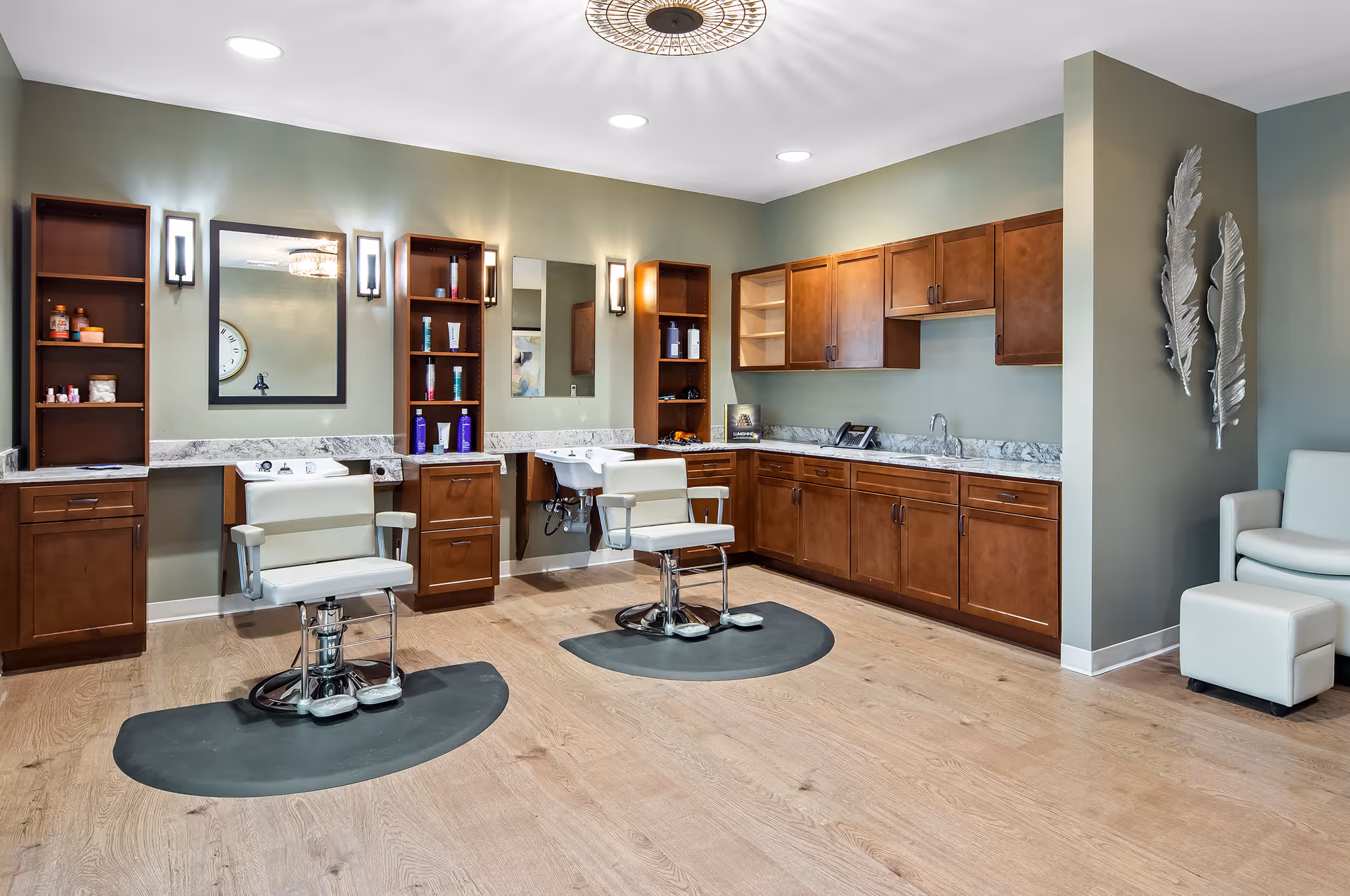 Interior view of a salon area with two white salon chairs on black mats in front of mirrors and wooden shelves stocked with hair care products. The room has wooden cabinets along one wall, a sink, and light green walls with decorative silver feather wall art. There is also a white armchair and ottoman in the corner.