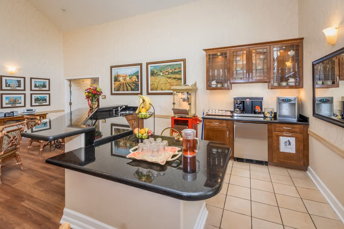 Interior view of a senior living facility kitchen area with a black granite countertop island. The island has a tiered fruit basket with bananas and apples, a tray with glasses, and a pitcher of iced tea. Behind the island, there are wooden cabinets with glass doors displaying dishes and glassware, a coffee machine, and a popcorn machine. The walls are decorated with framed landscape paintings and the floor is tiled near the kitchen and wooden in the adjacent area.