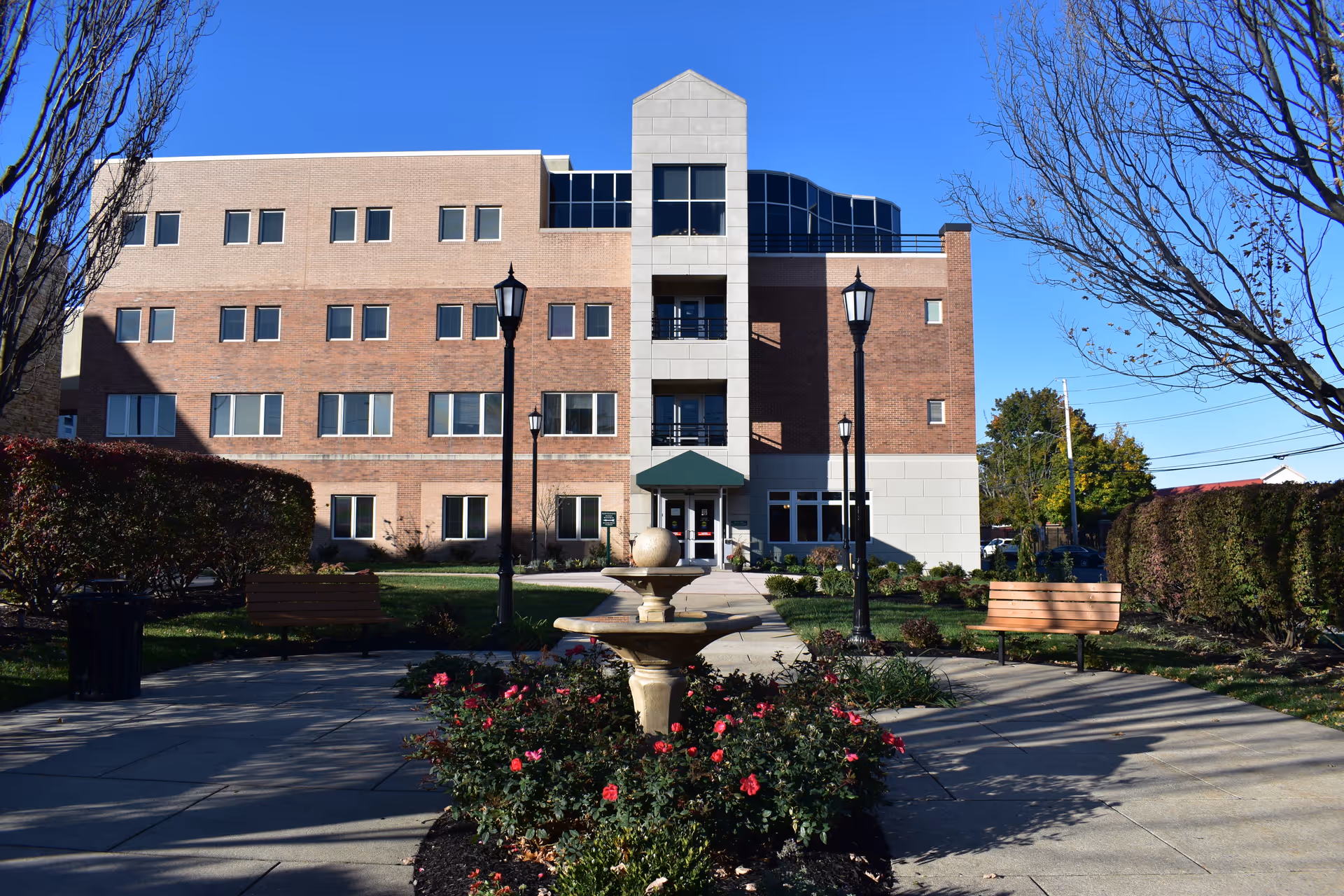 Front exterior of a multi-story brick senior living building with a central walkway, fountain, benches, and lamp posts.