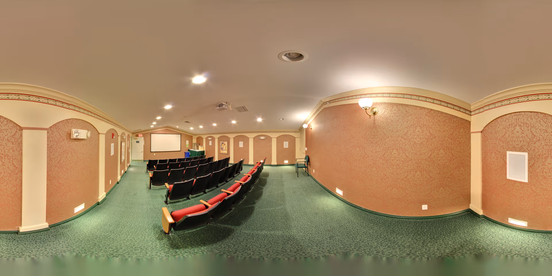 A small conference or presentation room with rows of black and red cushioned chairs facing a projection screen and a table with a green tablecloth. The room has patterned wallpaper in a warm tone, green carpet, and ceiling lights.